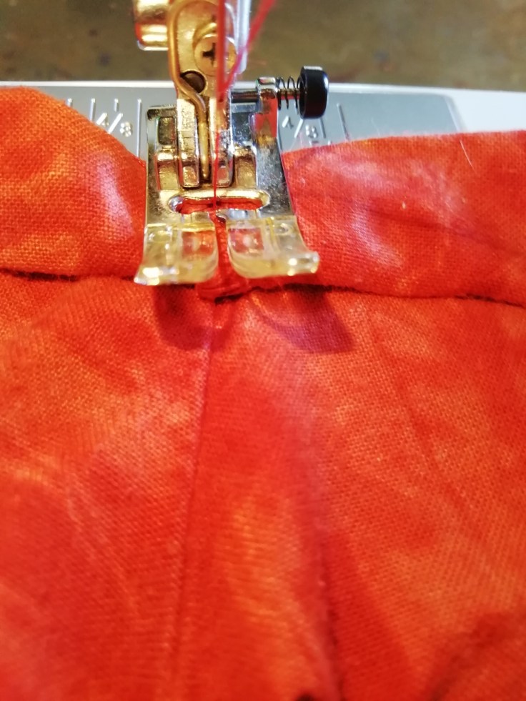 Tacking over the rolled sleeve hem. A sewing machine foot holds down the hem while the needle straight stitches a row across the roll.
